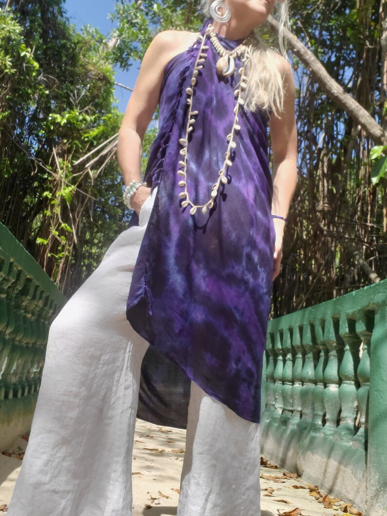 May include: A woman wearing a long, purple and blue tie-dye dress with a white shell necklace and white pants.