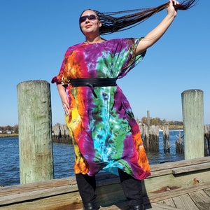 May include: A woman wearing a long, colorful tie-dye dress with a black belt. She is standing on a wooden dock with water in the background.