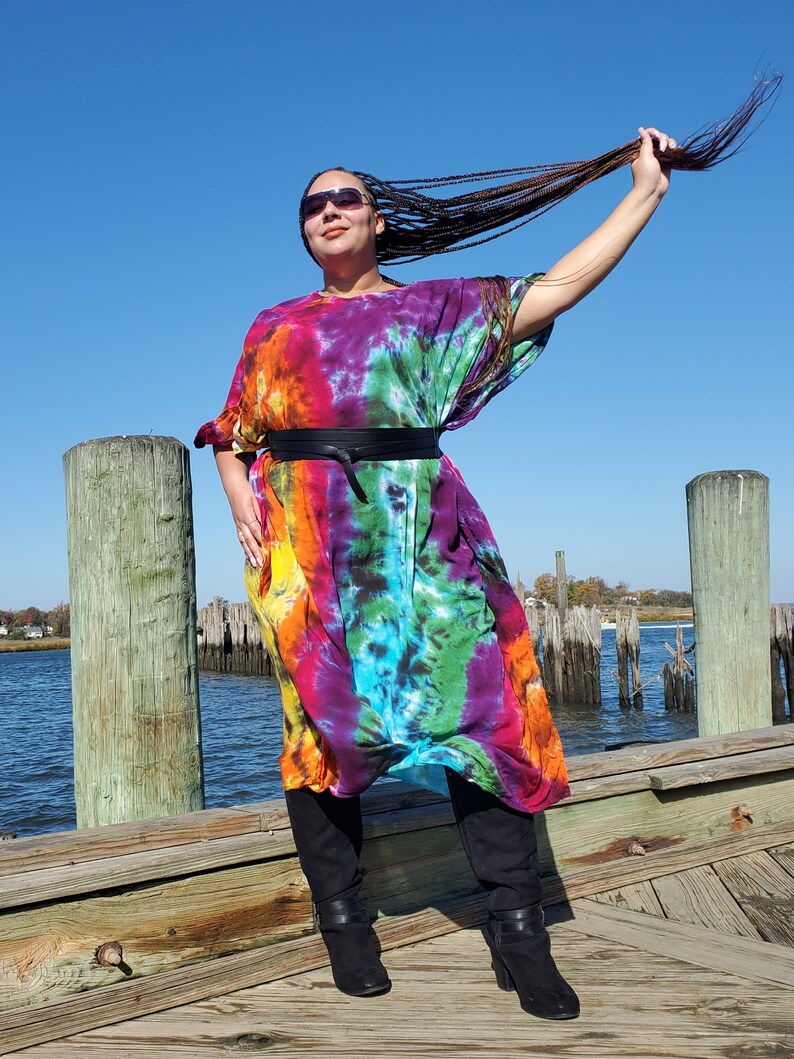 May include: A woman wearing a long, colorful tie-dye dress with a black belt. She is standing on a wooden dock with water in the background.