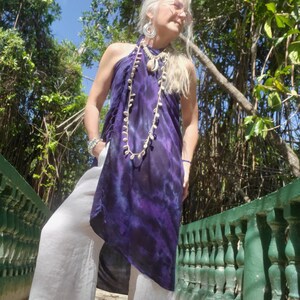 May include: A woman wearing a purple and white tie-dye halter top with a shell necklace and white pants. She is standing on a path with a green railing.