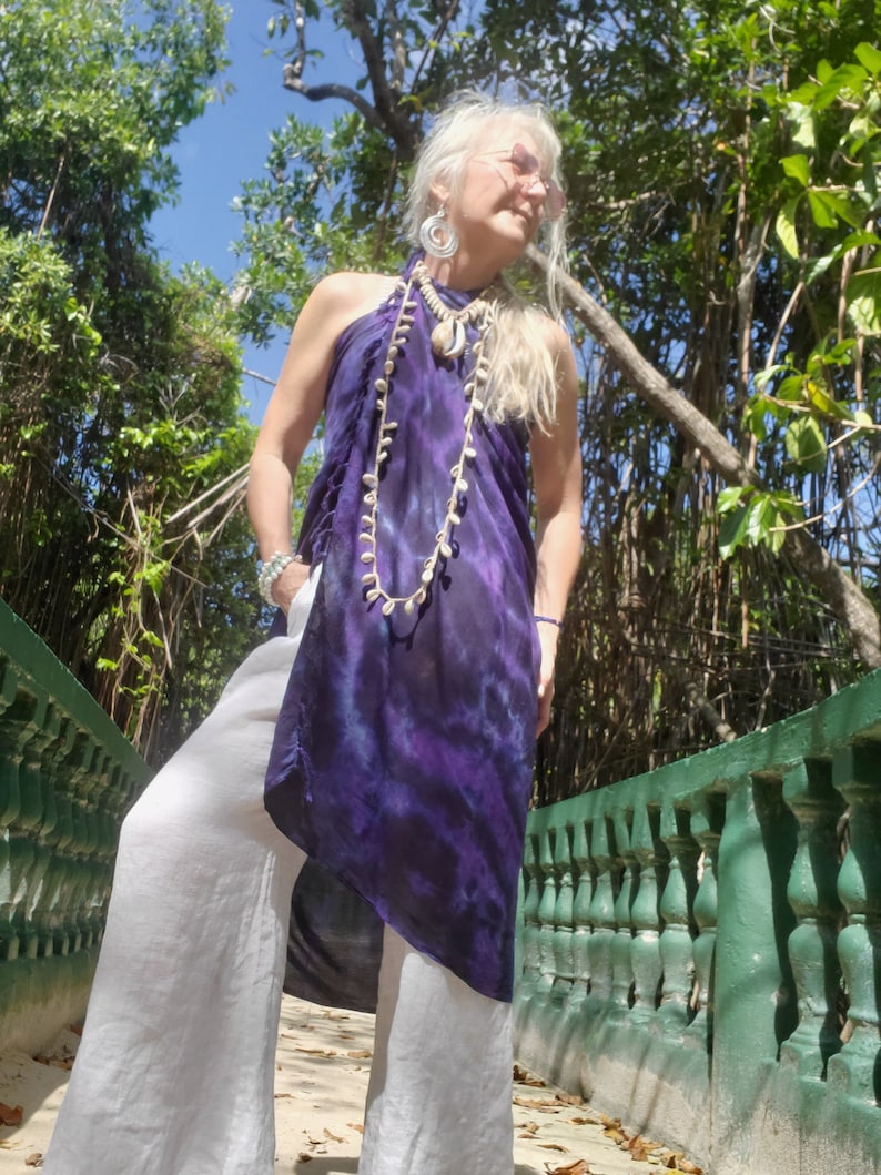 May include: A woman wearing a purple and white tie-dye halter top with a shell necklace and white pants. She is standing on a path with a green railing.
