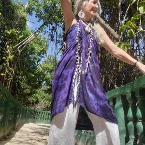 May include: A woman wearing a purple and white tie-dye sarong with a white shell necklace and white pants. She is standing on a walkway with a green railing.