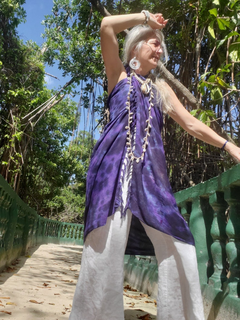 May include: A woman wearing a purple and white tie-dye sarong with a white shell necklace and white pants. She is standing on a walkway with a green railing.