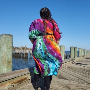 May include: A person wearing a long, colorful tie-dye dress with a black belt and black boots. The person is standing on a wooden dock overlooking a body of water.