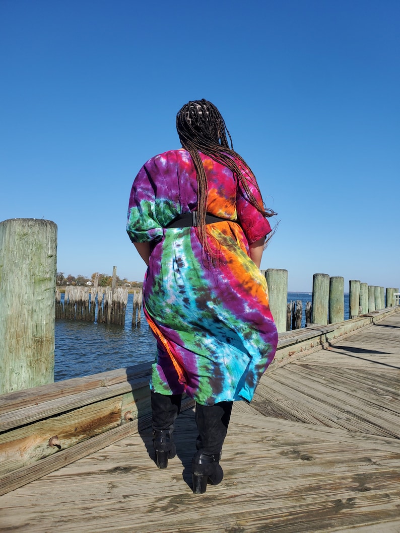 May include: A person wearing a long, colorful tie-dye dress with a black belt and black boots. The person is standing on a wooden dock overlooking a body of water.
