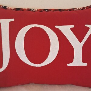 May include: Red throw pillow with white embroidered text "JOY" on the front. The pillow has a black and red plaid trim with white snowflakes.