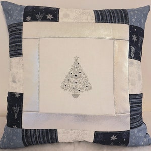 May include: A white throw pillow with a silver embroidered Christmas tree design. The pillow is made of patchwork fabric in shades of blue and white.