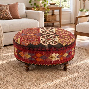 May include: A round ottoman featuring a multi-colored woven textile with geometric patterns in shades of red, orange, black, and cream. The piece is supported by four short, dark brown turned wooden legs, providing a stable base for the circular seat.