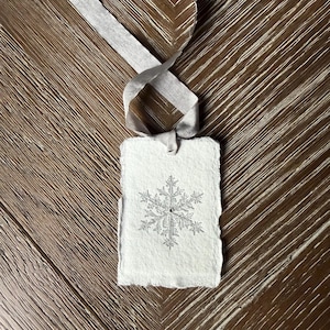 Könnte beinhalten: Ein rechteckiges, cremefarbenes Geschenk-Etikett mit einem silbernen Schneeflocken-Design und einem grauen Band. Das Etikett hat einen gezackten Rand und ist mit einem grauen Band gebunden. Der Hintergrund ist eine braune Holzoberfläche.