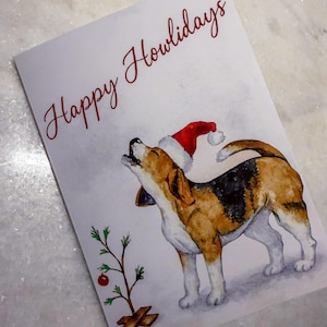 May include: A greeting card with a watercolor illustration of a beagle wearing a Santa hat, howling upwards. The card reads "Happy Howlidays" in red script. A small Christmas tree is on the left.