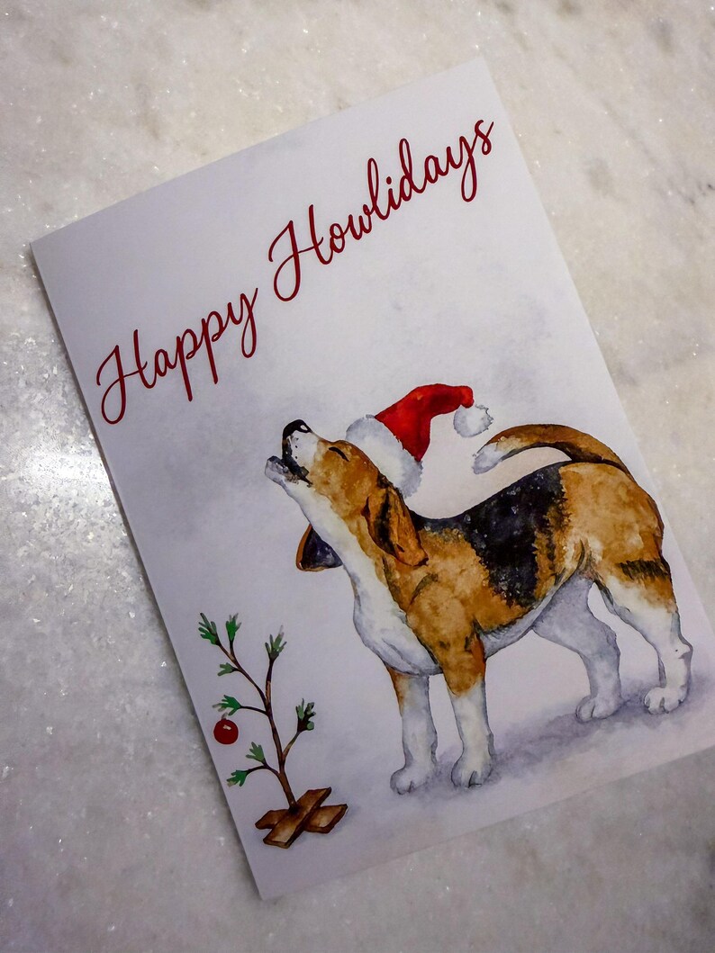May include: A greeting card with a watercolor illustration of a beagle wearing a Santa hat, howling upwards. The card reads "Happy Howlidays" in red script. A small Christmas tree is on the left.