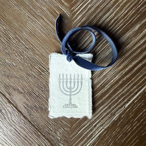 Könnte beinhalten: Ein kleines, rechteckiges Papieretikett mit einer grauen Menora-Illustration und einem dunkelblauen Samtband. Das Etikett hat einen Büttenrand und ist mit einer Schleife aus Band gebunden. Die Menora hat sieben Kerzenhalter. Der Hintergrund ist eine Holzoberfläche.