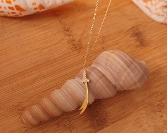 May include: A gold chain necklace with a diamond-encrusted sword pendant. The pendant is a curved sword with a pointed tip. The necklace is displayed on a seashell.