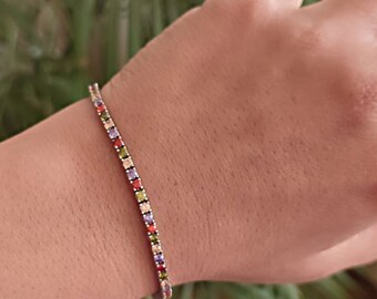 Può includere: Un braccialetto d'argento con una catena sottile e pietre quadrate colorate. Le pietre sono disposte in un motivo ripetitivo di rosso, verde, giallo e viola.