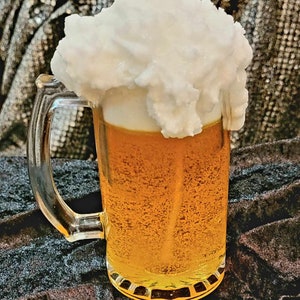 May include: A glass beer mug with a white foamy head, resembling a cloud, on top. The mug is filled with a golden yellow liquid.