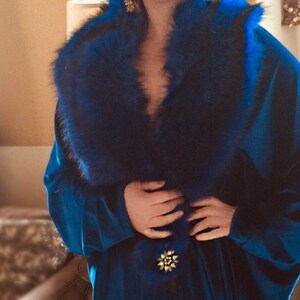 May include: A long, flowing, royal blue velvet robe with a plush, dark blue fur collar. The robe is secured with a gold and pearl brooch at the waist. The robe's design suggests a vintage or luxury aesthetic.
