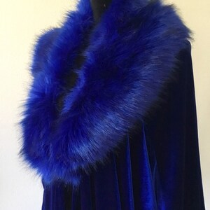 May include: A royal blue velvet cape with a matching faux fur collar. The plush collar is a vibrant blue, contrasting with the smooth velvet fabric. The cape is displayed on a black mannequin.