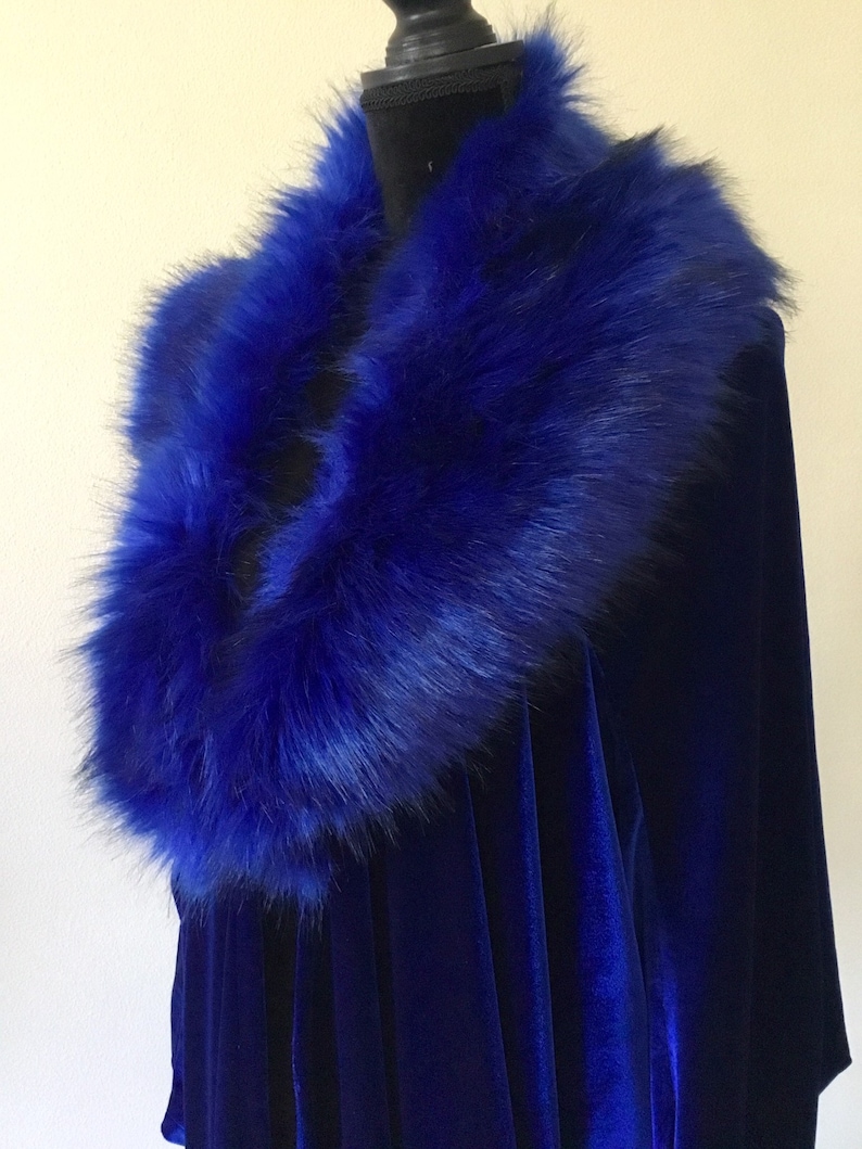 May include: A royal blue velvet cape with a matching faux fur collar. The plush collar is a vibrant blue, contrasting with the smooth velvet fabric. The cape is displayed on a black mannequin.