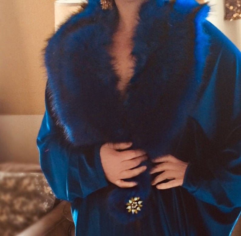 May include: A long, flowing, royal blue velvet robe with a plush, dark blue fur collar. The robe is secured with a gold and pearl brooch at the waist. The robe's design suggests a vintage or luxury aesthetic.