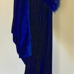 May include: A long, royal blue velvet cloak with a matching faux fur collar. The cloak is displayed on a black mannequin, showcasing its flowing design and rich color.