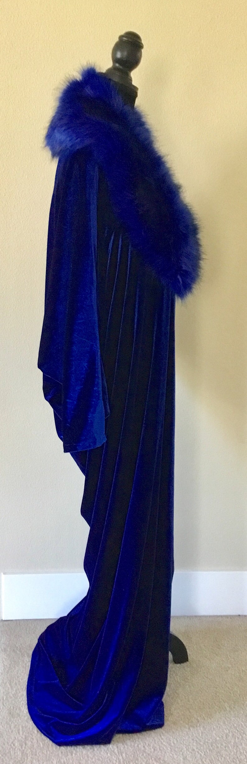 May include: A long, royal blue velvet cloak with a matching faux fur collar. The cloak is displayed on a black mannequin, showcasing its flowing design and rich color.