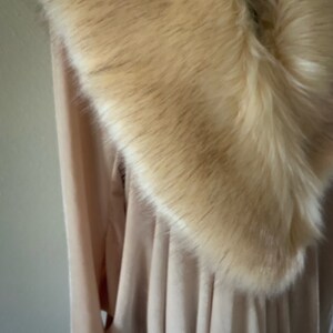 May include: A light beige long-sleeved garment featuring a wide, plush faux fur collar in a warm cream tone. The fabric of the garment appears soft and draped, with vertical folds visible beneath the thick, textured fur trim around the neckline.