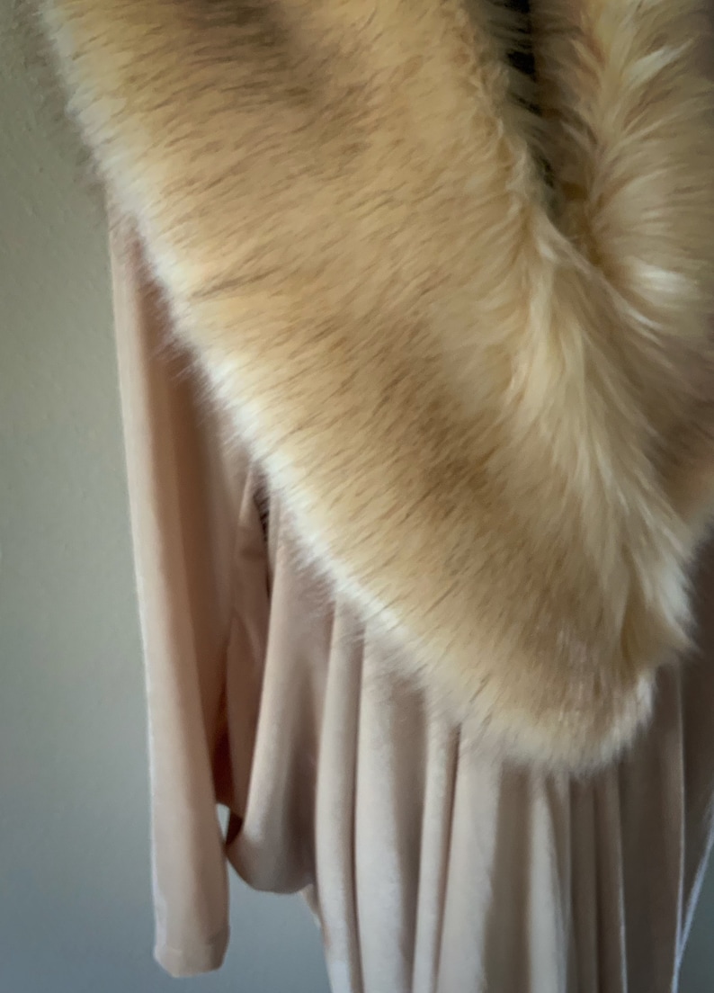 May include: A light beige long-sleeved garment featuring a wide, plush faux fur collar in a warm cream tone. The fabric of the garment appears soft and draped, with vertical folds visible beneath the thick, textured fur trim around the neckline.