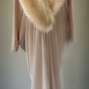 May include: A floor-length, light beige, flowing garment featuring a wide, plush, tan faux fur collar that drapes around the neckline. The fabric is soft and semi-sheer, falling in vertical folds from the shoulders down to the hemline.