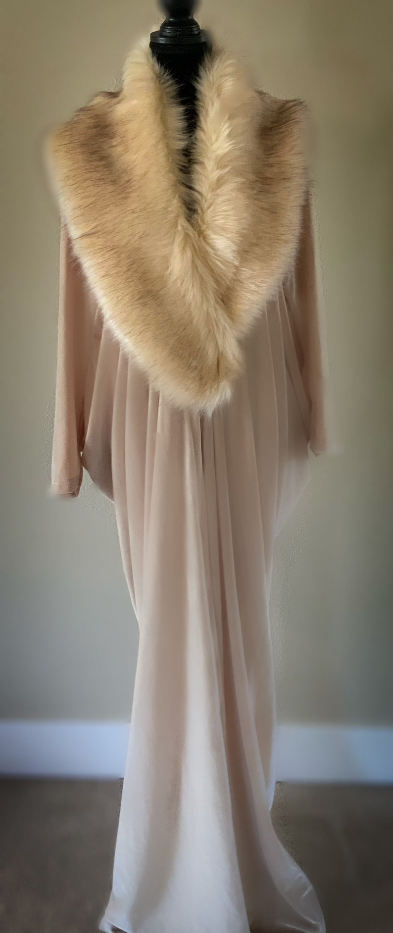 May include: A floor-length, light beige, flowing garment featuring a wide, plush, tan faux fur collar that drapes around the neckline. The fabric is soft and semi-sheer, falling in vertical folds from the shoulders down to the hemline.
