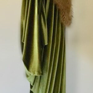 Puede incluir: Una larga t&uacute;nica de terciopelo verde con un cuello de piel marr&oacute;n. La t&uacute;nica est&aacute; drapeada sobre un maniqu&iacute; y tiene un dise&ntilde;o fluido y elegante.