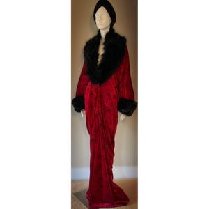 May include: A long, red velvet robe with black faux fur trim on the collar and cuffs. The robe is displayed on a mannequin, paired with a black turban. The robe has a flowing design and a slight train.