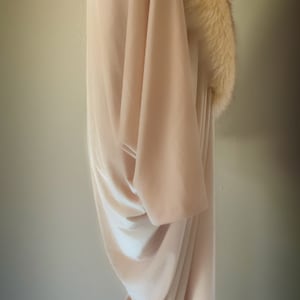 May include: A floor-length, light beige velvet gown featuring a wide, plush, cream-coloured faux fur collar that drapes around the neckline and shoulders. The garment has long, loose sleeves and a fluid, draped silhouette that falls to the floor.