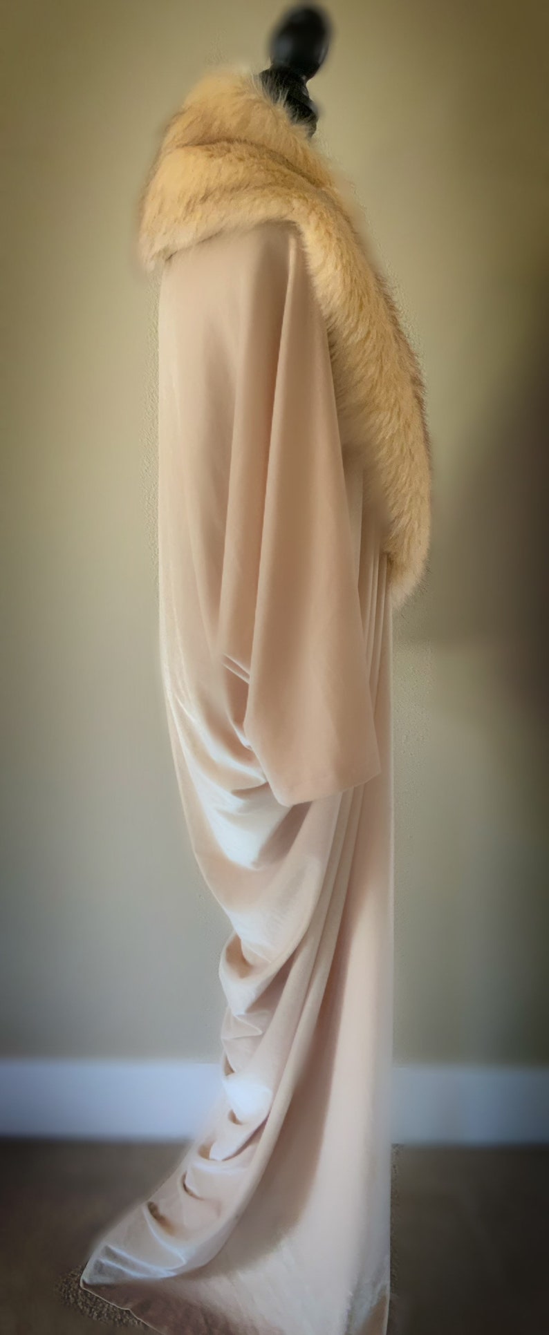 May include: A floor-length, light beige velvet gown featuring a wide, plush, cream-coloured faux fur collar that drapes around the neckline and shoulders. The garment has long, loose sleeves and a fluid, draped silhouette that falls to the floor.