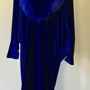 May include: A long, flowing, royal blue velvet gown with a matching faux fur collar. The dress is displayed on a black mannequin, showcasing its elegant drape and design. The gown's sleeves are long and the hemline reaches the floor.