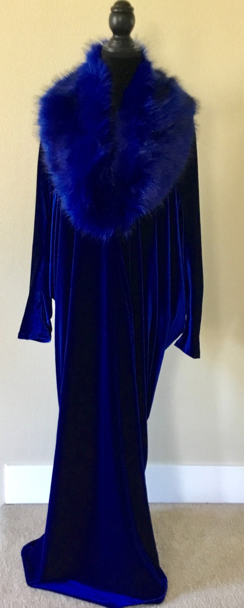 May include: A long, flowing, royal blue velvet gown with a matching faux fur collar. The dress is displayed on a black mannequin, showcasing its elegant drape and design. The gown's sleeves are long and the hemline reaches the floor.