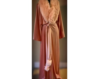 May include: A long, peach-coloured velvet robe with a faux fur collar and matching belt. The robe has wide sleeves and a flowing design. The faux fur collar is a lighter shade of peach. The robe is displayed on a black mannequin.
