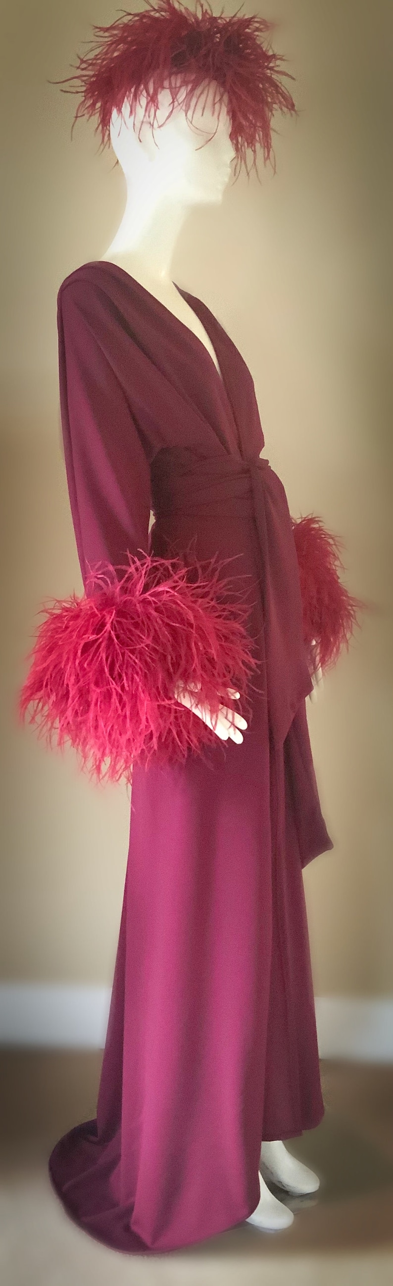 May include: A long, burgundy robe with a deep V-neckline and feather trim on the cuffs and collar. The robe is made of a soft, flowing fabric and has a tie belt.