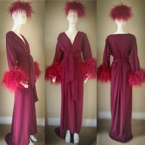 May include: A long, burgundy robe with a V-neckline and long sleeves. The robe has a sash tie at the waist and feather trim on the cuffs and collar.
