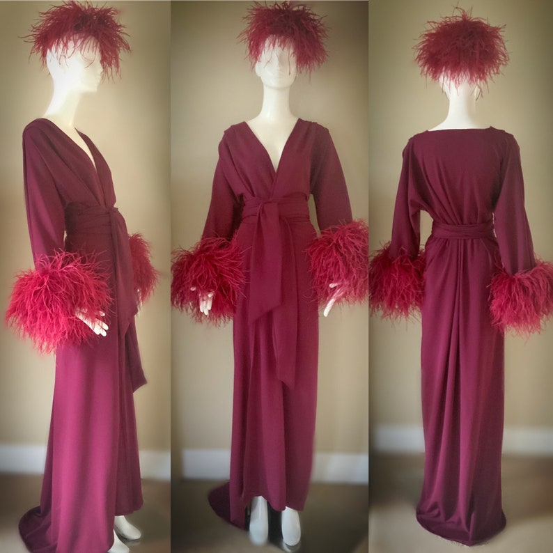 May include: A long, burgundy robe with a V-neckline and long sleeves. The robe has a sash tie at the waist and feather trim on the cuffs and collar.
