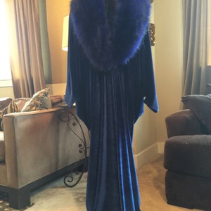 May include: A long, flowing, navy blue velvet gown with a large, fluffy, royal blue fur collar. The dress is displayed on a mannequin, showcasing its elegant drape and design. The gown's train pools on the floor.