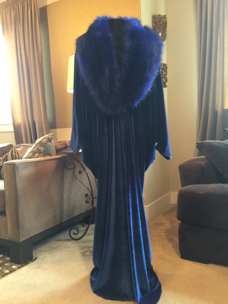 May include: A long, flowing, navy blue velvet gown with a large, fluffy, royal blue fur collar. The dress is displayed on a mannequin, showcasing its elegant drape and design. The gown's train pools on the floor.