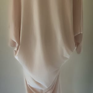 May include: A floor-length, light beige dress featuring a draped, loose-fitting silhouette with wide sleeves and a high neckline adorned with a thick, textured faux fur collar in shades of cream, tan, and dark brown, creating a soft, layered appearance.