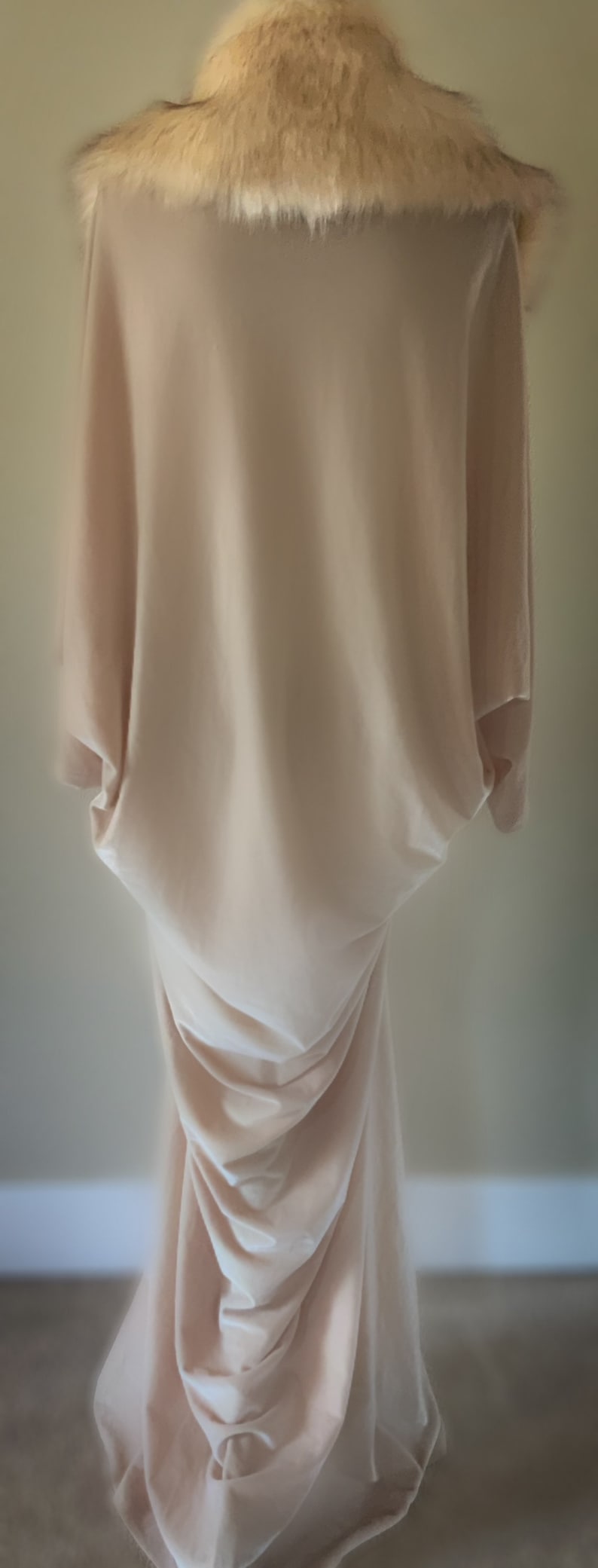 May include: A floor-length, light beige dress featuring a draped, loose-fitting silhouette with wide sleeves and a high neckline adorned with a thick, textured faux fur collar in shades of cream, tan, and dark brown, creating a soft, layered appearance.