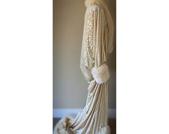 May include: A cream-coloured, floor-length robe with a floral pattern, displayed on a mannequin. The robe features a faux fur collar, cuffs, and hem. The fabric has a subtle sheen, and the overall design suggests a vintage or boudoir aesthetic.