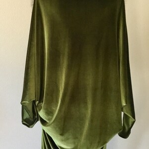 Puede incluir: Una larga t&uacute;nica de terciopelo verde con un cuello de piel sint&eacute;tica marr&oacute;n. La t&uacute;nica est&aacute; drapeada sobre un maniqu&iacute; y tiene una silueta fluida y elegante.