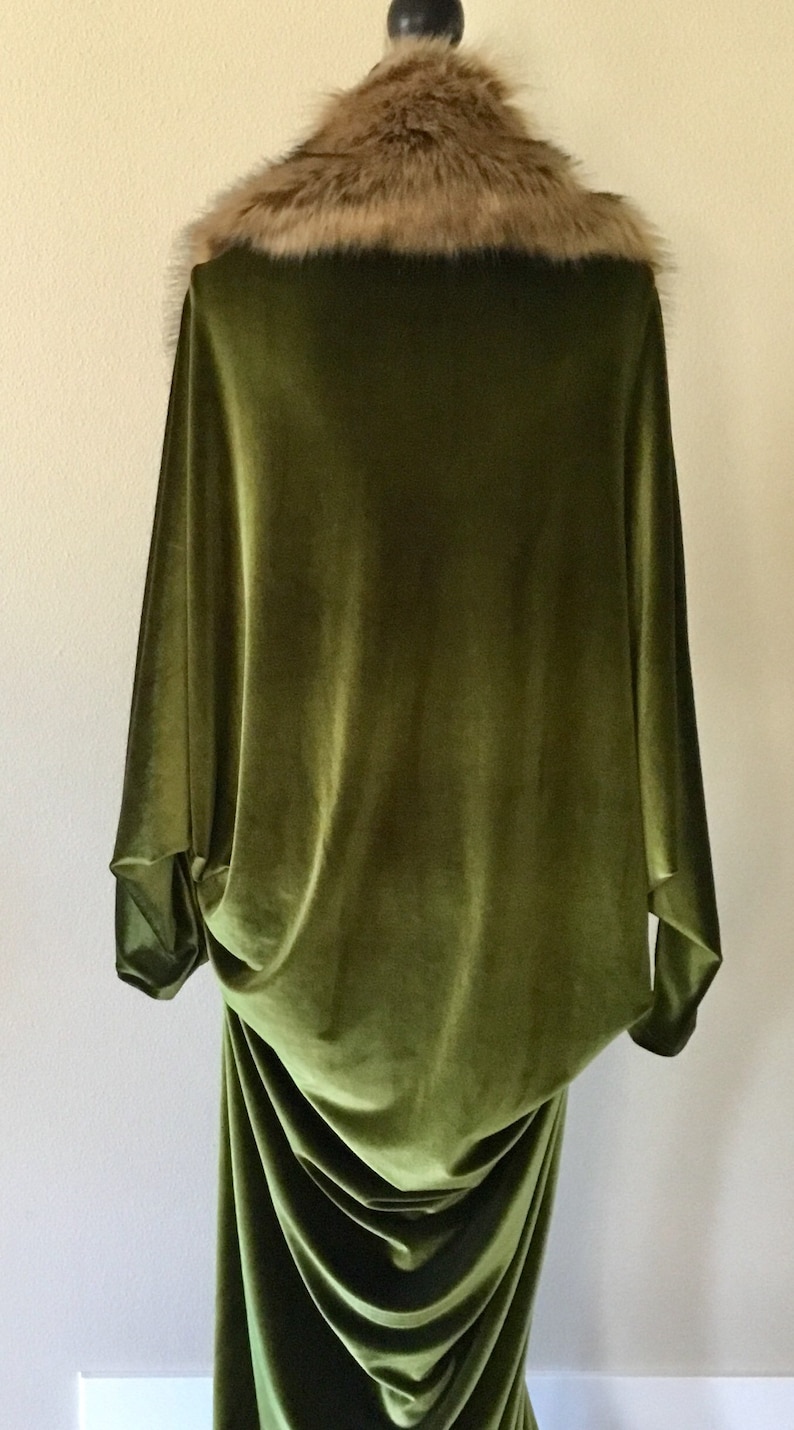 Puede incluir: Una larga t&uacute;nica de terciopelo verde con un cuello de piel sint&eacute;tica marr&oacute;n. La t&uacute;nica est&aacute; drapeada sobre un maniqu&iacute; y tiene una silueta fluida y elegante.