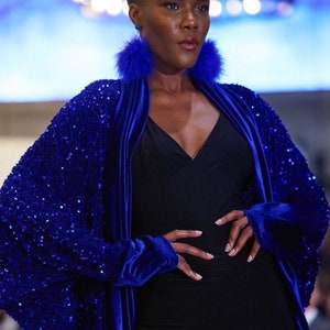 May include: A model wearing a black dress and a royal blue sequined jacket with velvet trim. The jacket has wide sleeves and is open in the front. The model is also wearing blue feather earrings.