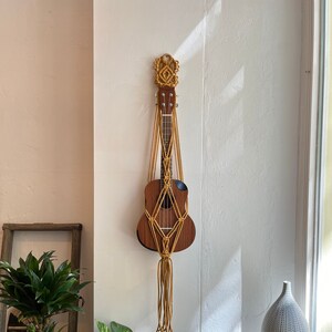 Może przedstawiać: Ukelele wiszące na białej ścianie z makramą wykonaną z żółtego sznurka. Ukelele jest brązowe z czarnym pickguardem.