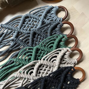 Puede incluir: Primer plano de cinco colgadores de plantas de macramé en varios colores: gris, verde, beige y negro. Cada colgador tiene un anillo de madera en la parte superior. Los colgadores están hechos de cuerda de algodón tejida, de estilo bohemio.