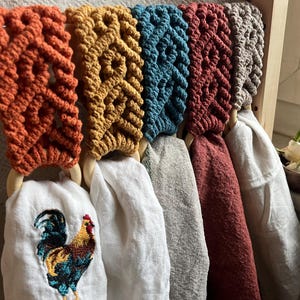 May include: Six colourful macrame towel holders with wooden rings, hanging on a wooden rack. Each holder is a different colour: orange, yellow, teal, red, brown, and grey. The towel holders are hanging over white, beige, red, and grey towels. A rooster is embroidered on one of the towels.